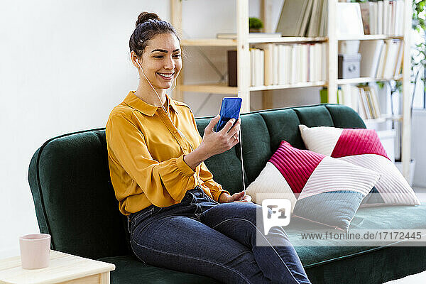 Woman with in-ear headphones taking selfie through mobile phone while sitting at home
