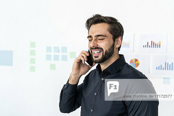 Smiling businessman talking on smart phone with eyes closed against wall in office