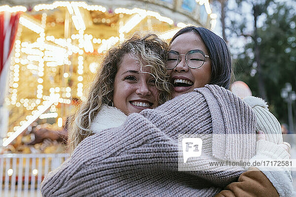 Laughing female friends embracing against illuminated carousel