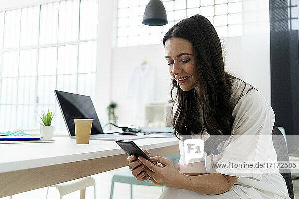 Lächelnde junge Ärztin mit Smartphone am Schreibtisch in der Klinik
