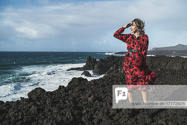 Junge Frau in rotem Kleid betrachtet die Aussicht  während sie auf einem Berg in Los Hervideros  Lanzarote  Spanien  steht