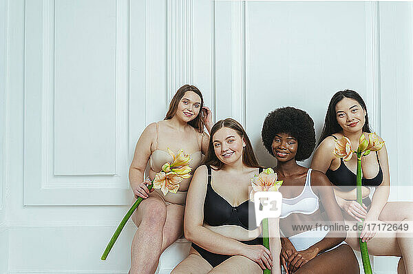 Lächelnde multiethnische Gruppe weiblicher Modelle in Unterwäsche  die Kunstblumen vor einer weißen Wand halten