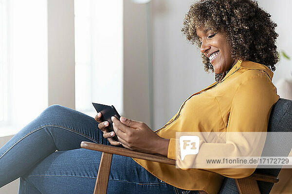 Frau lächelt  während sie ein digitales Tablet benutzt und sich auf einem Sessel zu Hause entspannt