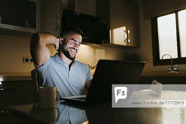 Lächelnder Geschäftsmann mit Händen hinter dem Kopf bei der Arbeit am Laptop am Esstisch zu Hause