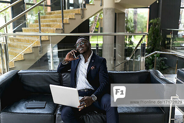 Gutaussehender männlicher Geschäftsmann mit Laptop  der auf einem Sofa im Hotel sitzt und telefoniert
