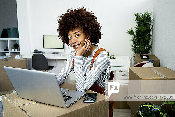 Lächelnde junge Frau mit Hand am Kinn vor einem Laptop im Wohnzimmer ihres neuen Hauses