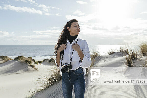 Young woman with vintage camera standing on sand dunes against sky at beach