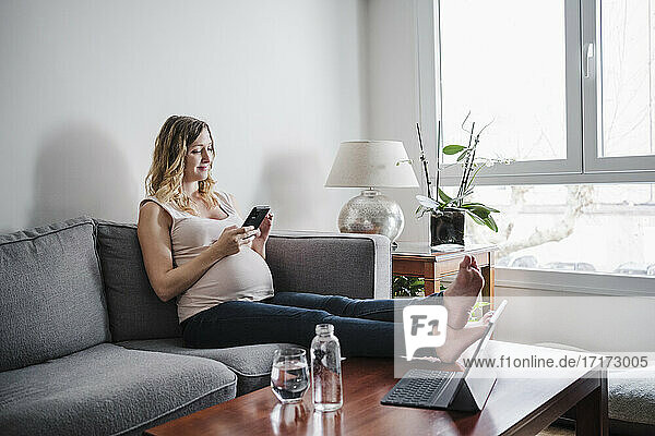 Schwangere Frau benutzt ihr Smartphone  während sie zu Hause auf dem Sofa sitzt