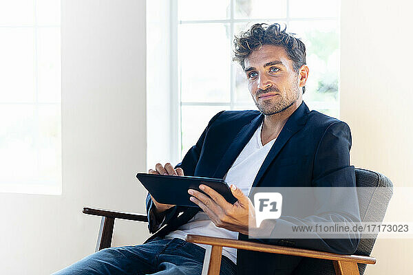 Nachdenklicher Geschäftsmann mit digitalem Tablet  der wegschaut  während er zu Hause auf einem Sessel sitzt