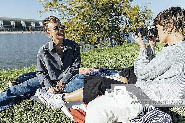 Junge Frau fotografiert einen lächelnden Freund durch die Kamera  während sie im Park sitzt