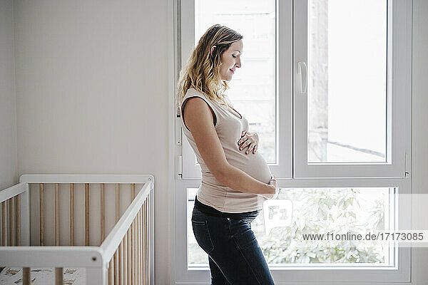 Schwangere Frau mit Händen auf dem Bauch nachdenklich  während sie zu Hause am Fenster steht