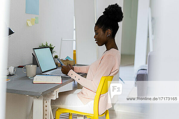 Young businesswoman with laptop using mobile phone while sitting at home office