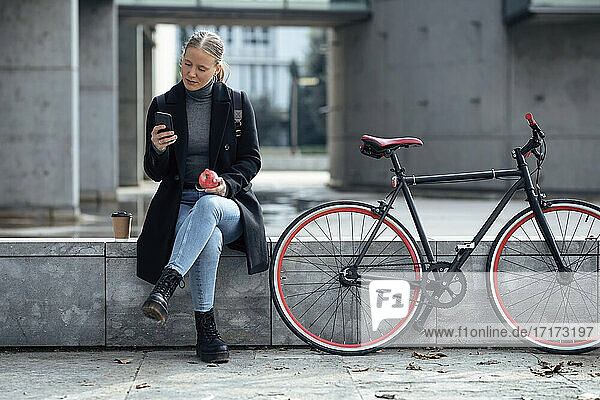 Young woman using mobile phone while sitting on retaining wall by bicycle