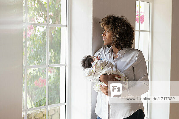 Thoughtful woman with baby looking away while standing by window at home