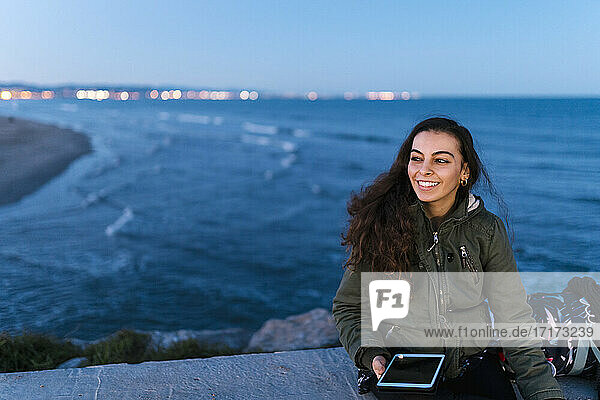 Smiling woman with digital tablet looking away while sitting on retaining wall against sea