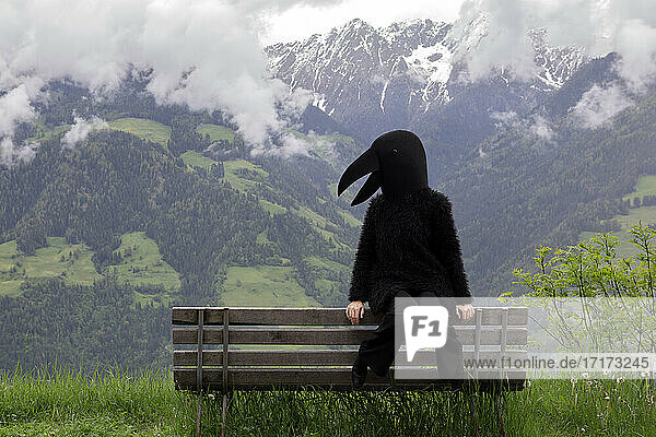 Female in crow costume sitting on bench against mountain range