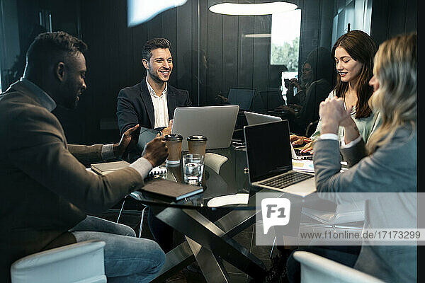 Smiling businessman with laptop having discussion with team while sitting in meeting at office