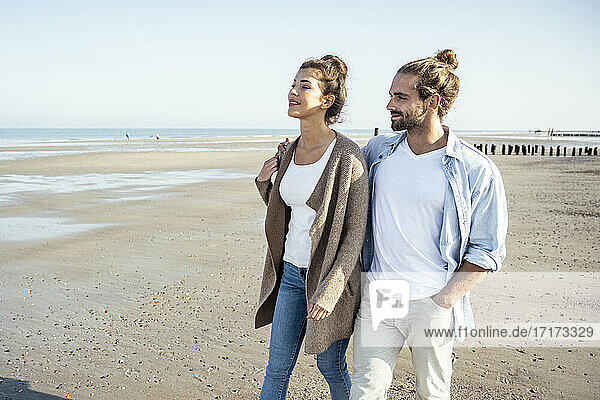 Smiling girlfriend and boyfriend looking away while walking on beach during weekend
