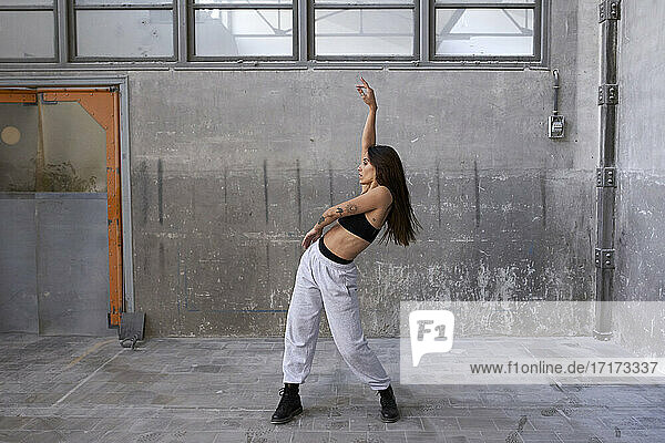 Female dancer dancing with arm raised against gray wall in abandoned factory