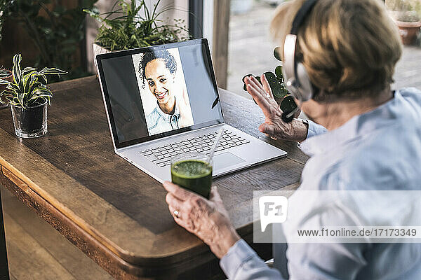 Großmutter winkt ihrer Enkelin per Videoanruf über den Laptop zu  während sie zu Hause Saft trinkt