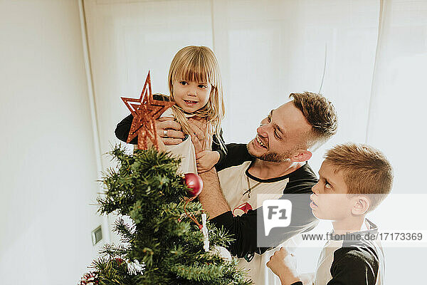 Father showing Christmas tree decoration while carrying daughter by son against wall at home