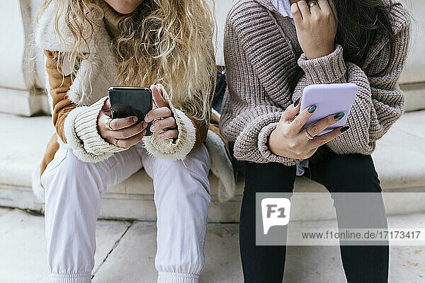 Female friends in warm clothing using mobile phone while sitting on steps