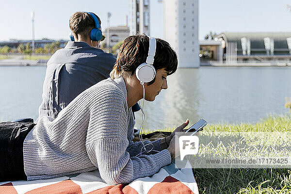 Frau mit Kopfhörern  die ein Mobiltelefon benutzt  während sie auf einer Decke neben einem Freund am Fluss liegt