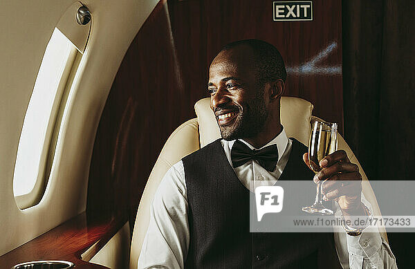 Lächelnder männlicher Unternehmer mit Champagner in der Hand  der aus dem Fenster eines Flugzeugs schaut