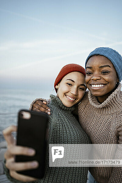 Junge Frauen mit Strickmütze  die ein Selfie mit ihrem Mobiltelefon machen  während sie vor dem Himmel stehen