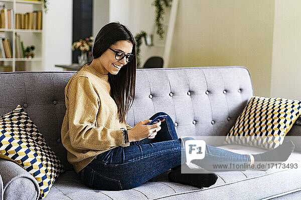 Smiling young woman using smart phone while sitting on sofa at home