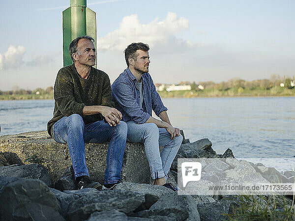 Thoughtful father and son looking away while sitting on rock against sky