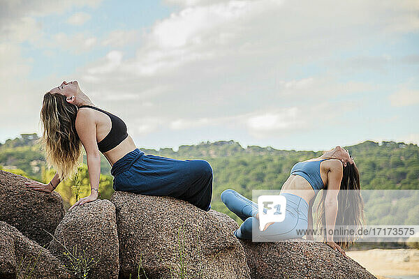 Flexible young female instructor and young woman exercising on rock against sky
