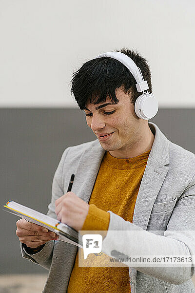 Lächelnder Mann mit Kopfhörern  der in ein Buch schreibt  während er an der Wand steht