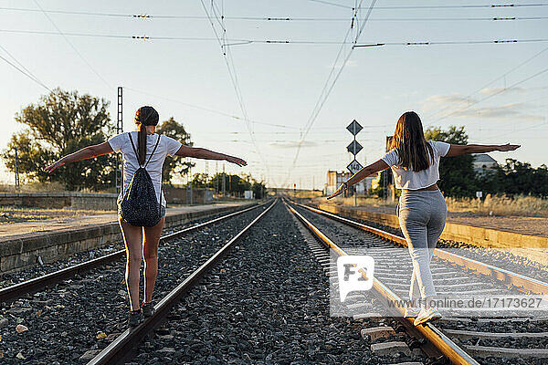 Freundinnen mit ausgestreckten Armen gehen auf Eisenbahnschienen gegen den Himmel bei Sonnenuntergang