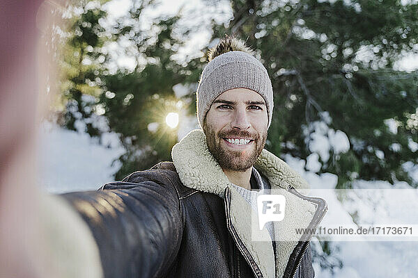 Glücklicher junger Mann nimmt Selfie gegen Bäume bei kaltem Wetter