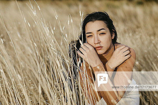 Serious young woman sitting amidst grass in field during sunset