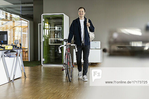 Junger Geschäftsmann hält Tasche  während er mit dem Fahrrad im Büro spazieren geht