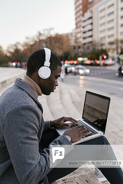 Young man wearing headphones working on laptop while sitting in city