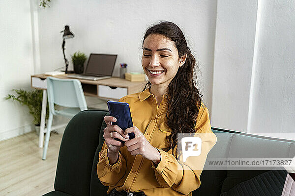 Young woman smiling while using mobile phone sitting on sofa at home