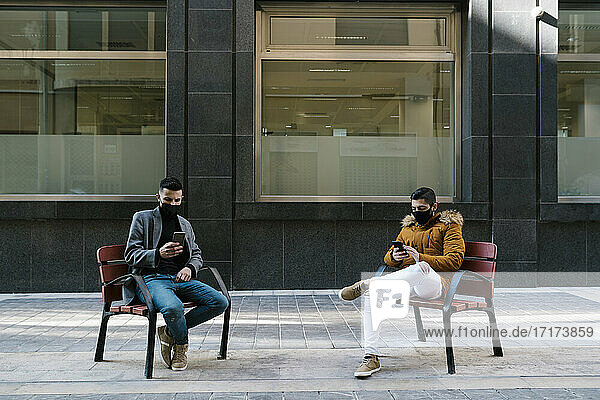 Junge Männer mit Gesichtsmaske benutzen Mobiltelefone  während sie auf einem Stuhl in der Stadt sitzen