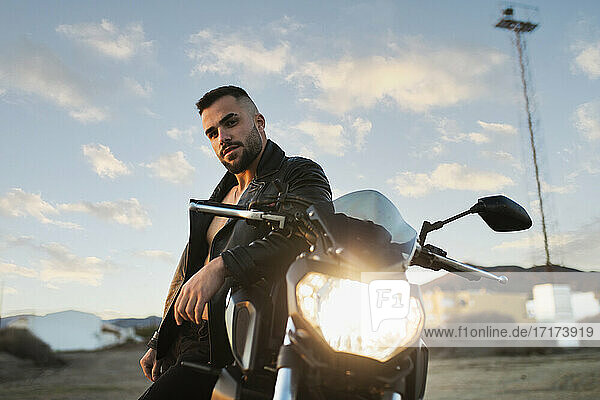Male biker with cool attitude leaning on motorcycle against sky
