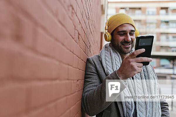 Mann mit Kopfhörern  der lächelnd sein Handy benutzt und sich an die Wand lehnt