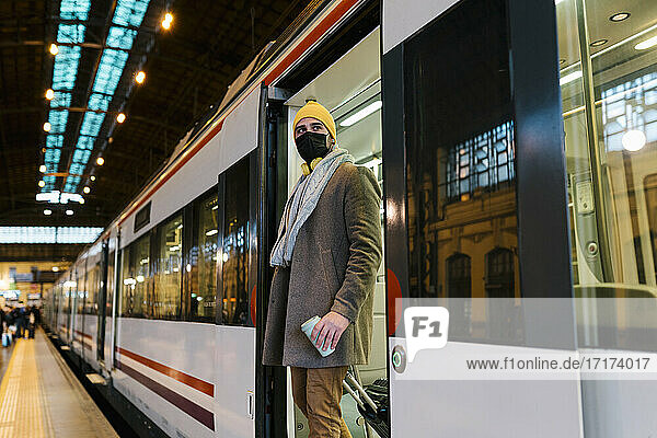 Mittlerer erwachsener Mann mit Gesichtsmaske  der mit Kaffeetasse und Gepäck am Bahnhof aus dem Zug steigt