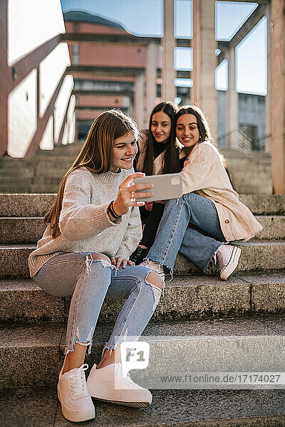Lächelndes Teenager-Mädchen  das ein Selfie mit Freunden macht  während es auf einer Treppe in der Stadt sitzt