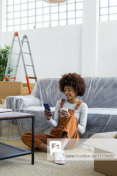 Afro-Frau mit Kaffeetasse  die ihr Smartphone benutzt  während sie auf dem Sofa in ihrem neuen Zuhause sitzt