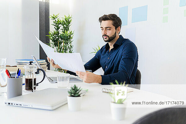 Male entrepreneur looking at documents while sitting against wall in office