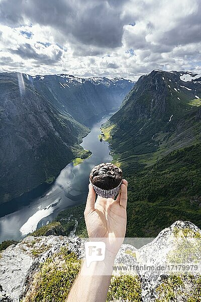 Rest with view  hand holding muffin  summit reward  view from the top of Breiskrednosi  mountains and fjord  Nærøyfjord  Aurland  Norway  Europe