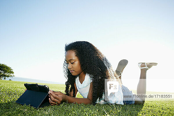 Neun Jahre altes  gemischtrassiges Mädchen liegt im Gras und schaut auf ein digitales Tablet.