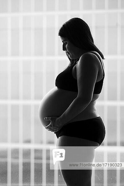 Seitenansicht einer schwangeren Frau  die im Studio steht und mit der Hand ihren Bauch hält