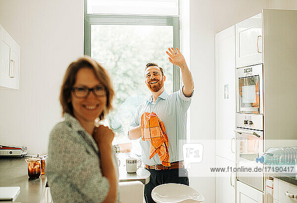 Ehepaar in der Küche  das Gäste empfängt. Mann lächelt und winkt mit der Hand